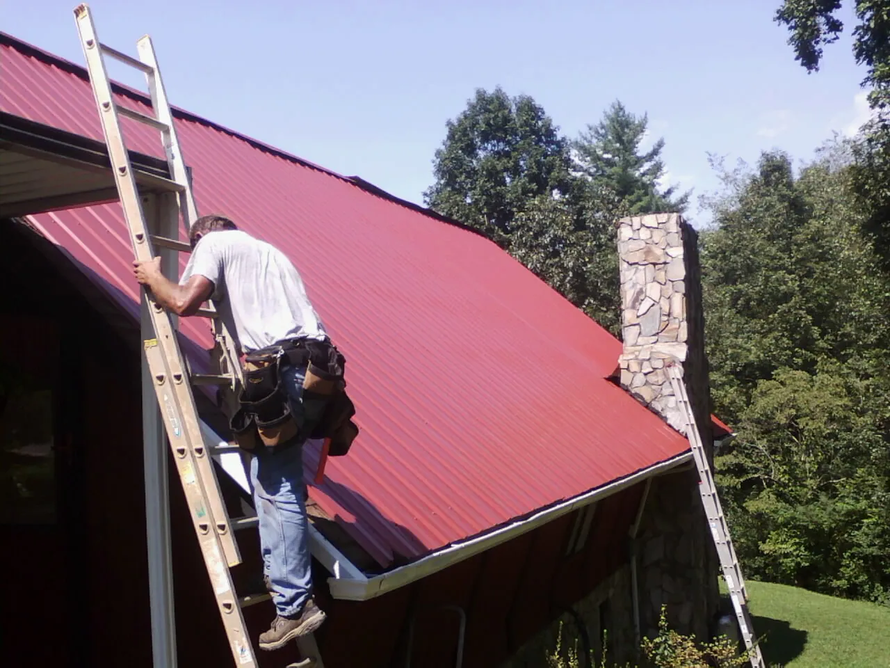 Roofer on a ladder beside a red metal roof and stone chimney — Soffit & Fascia Repair by Roofing Brothers in Ethelsville